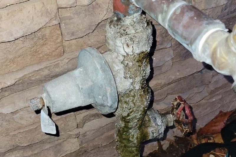 Old corroded hose bib and pressure regulator caked in mineral buildup against a stone wall
